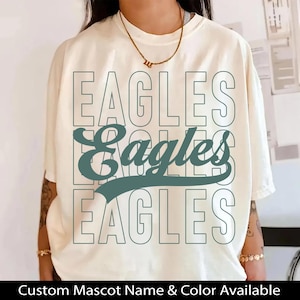 May include: Cream-colored t-shirt with the word "EAGLES" stacked three times in a teal font. The word "Eagles" is also written in a cursive font in the center. The shirt is a short-sleeved, casual style.