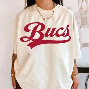 Op de afbeelding: Crèmekleurig T-shirt met het woord "Bucs" in een vet, rood, cursief lettertype over de voorkant. Het shirt heeft korte mouwen en een relaxte pasvorm. Een gouden ketting is zichtbaar bij de halslijn.