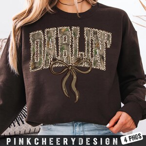 May include: A dark brown sweatshirt with the word "DARLIN" in a camouflage and leopard print design. A brown bow with glitter accents is below the text. The text "PINKCHEERYDESIGN 4 PNGS" is at the bottom.