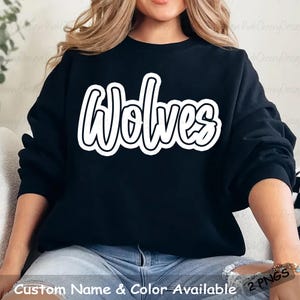 May include: Black sweatshirt with the word "Wolves" in large, white, outlined lettering across the chest. The text has a casual, handwritten style. The bottom of the image has the text "Custom Name & Color Available 2 PNGS".