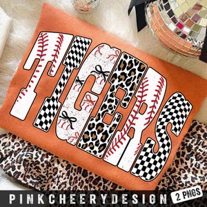May include: An orange t-shirt with the word "TIGERS" in a bold, decorative font. The letters feature baseball stitching, checkered patterns, and leopard print. The shirt is folded on a leopard print fabric.