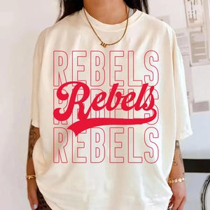 May include: Cream-colored t-shirt with the word "REBELS" in a stacked design, with the word "Rebels" in a cursive font in the middle. The text is in red. The shirt has short sleeves and a relaxed fit.