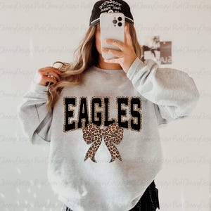 May include: A light grey sweatshirt featuring the word "EAGLES" in black block letters with a leopard print border. A leopard print bow is centered below the word. The sweatshirt is a long-sleeved crew neck.