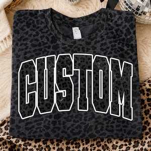 May include: A dark gray t-shirt with a leopard print pattern. The word "CUSTOM" is printed across the front in large, bold, white-outlined letters. The letters are filled with the same leopard print as the shirt.