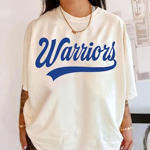 Puede incluir: Camiseta color crema con la palabra "Warriors" en negrita azul en la parte delantera. La palabra está arqueada sobre una pancarta azul. La camiseta tiene mangas cortas y un corte holgado.