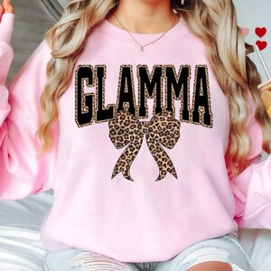 May include: A light pink sweatshirt with the word "GLAMMA" in large black letters with a leopard print fill. A leopard print bow is centered below the text. The sweatshirt is a casual style.