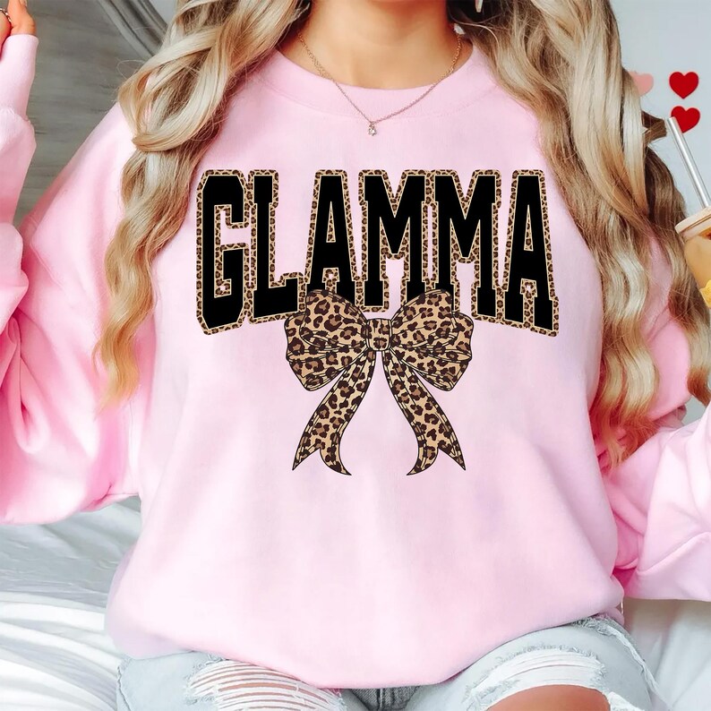 May include: A light pink sweatshirt with the word "GLAMMA" in large black letters with a leopard print fill. A leopard print bow is centered below the text. The sweatshirt is a casual style.