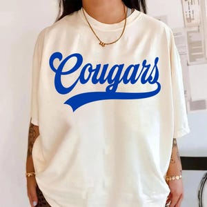 May include: Cream-colored t-shirt with the word "Cougars" in bold blue script across the front. The shirt has short sleeves and a relaxed fit. A gold necklace is visible at the neckline.