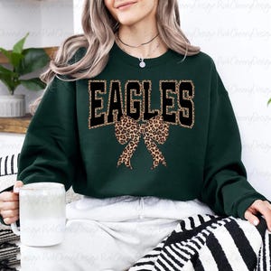 May include: A dark green sweatshirt featuring the word "EAGLES" in black block letters with a leopard print outline. A leopard print bow is positioned below the text. The sweatshirt has a crew neck.