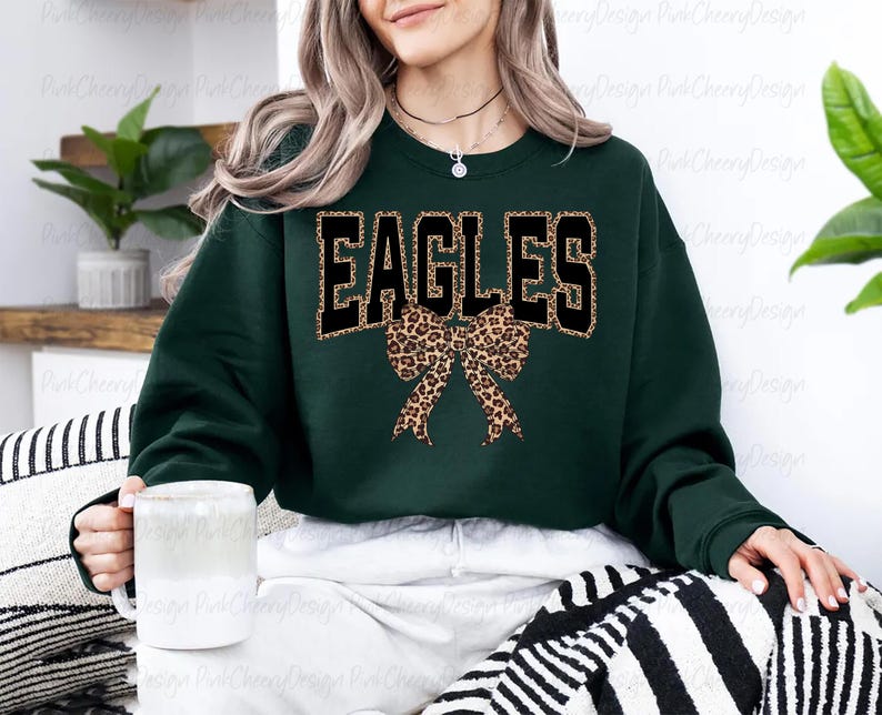 May include: A dark green sweatshirt featuring the word "EAGLES" in black block letters with a leopard print outline. A leopard print bow is positioned below the text. The sweatshirt has a crew neck.