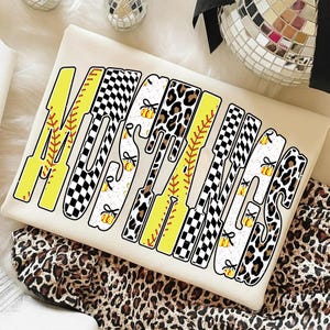 May include: Cream-colored t-shirt with the word "MAMAS" in a mix of yellow, black and white patterns. The letters are designed with baseball bat and softball elements, including stitching and a pumpkin graphic. The shirt is on a leopard print background.