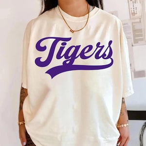 Könnte beinhalten: Cremefarbenes T-Shirt mit dem Wort "Tigers" in kräftiger, violetter Schrift auf der Vorderseite. Das Shirt hat kurze Ärmel und eine lockere Passform. Eine goldene Halskette ist am Ausschnitt zu sehen. Der Stil ist lässig und bequem.