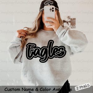 May include: A light gray sweatshirt with the word "Eagles" in a black and white design. The sweatshirt has long sleeves. The text "Custom Name & Color Available" is at the bottom.