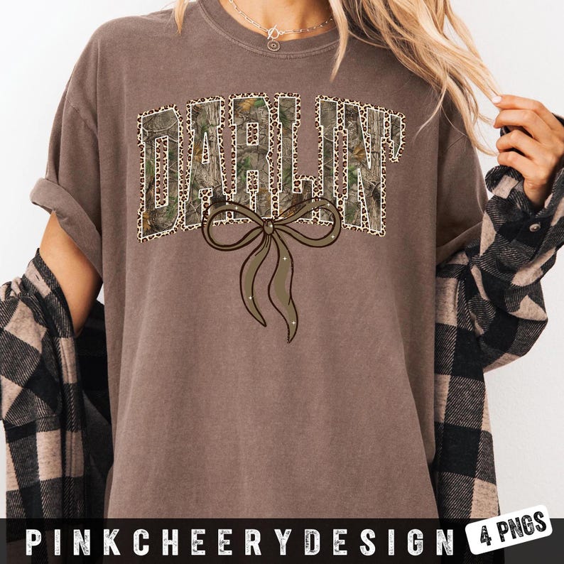 May include: A brown t-shirt with the word "DARLIN'" in a camouflage and leopard print design, with a brown bow graphic. The shirt has rolled-up sleeves and is part of a clothing collection.