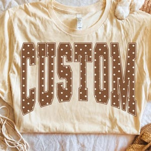 May include: A cream-colored short-sleeve t-shirt featuring the word CUSTOM printed in large, brown, varsity-style block letters. The letters are decorated with a repeating pattern of small white polka dots across the entire surface.