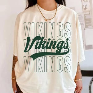 Può includere: T-shirt color crema con la scritta "Vikings" in un design a strati. Il testo superiore e inferiore è delineato in verde scuro, mentre il testo centrale è verde scuro pieno. Una maglietta casual per tutti i giorni.