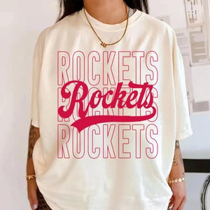 May include: Cream-colored t-shirt with the word "ROCKETS" in a stacked, red font. The word "Rockets" is repeated three times, with the middle word in a cursive style. The shirt has short sleeves and a relaxed fit.