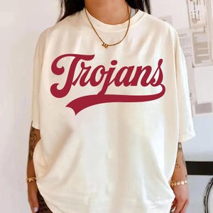 May include: Cream-colored t-shirt with the word "Trojans" in maroon script across the front. The shirt has short sleeves and a relaxed fit. A gold necklace is visible at the neckline.