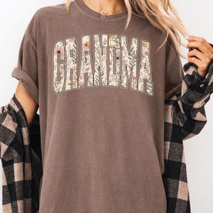 Op de afbeelding: Bruin t-shirt met het woord "GRANDMA" in een bloemenontwerp. De letters zijn gevuld met bloemen in roze, geel en groen. Het shirt heeft korte mouwen en een relaxte pasvorm.