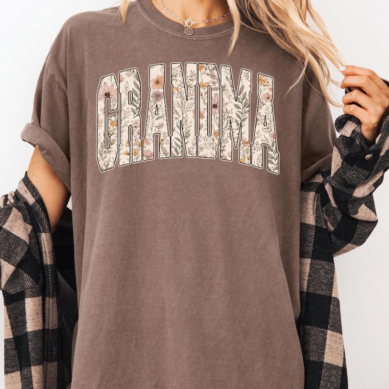 Op de afbeelding: Bruin t-shirt met het woord "GRANDMA" in een bloemenontwerp. De letters zijn gevuld met bloemen in roze, geel en groen. Het shirt heeft korte mouwen en een relaxte pasvorm.