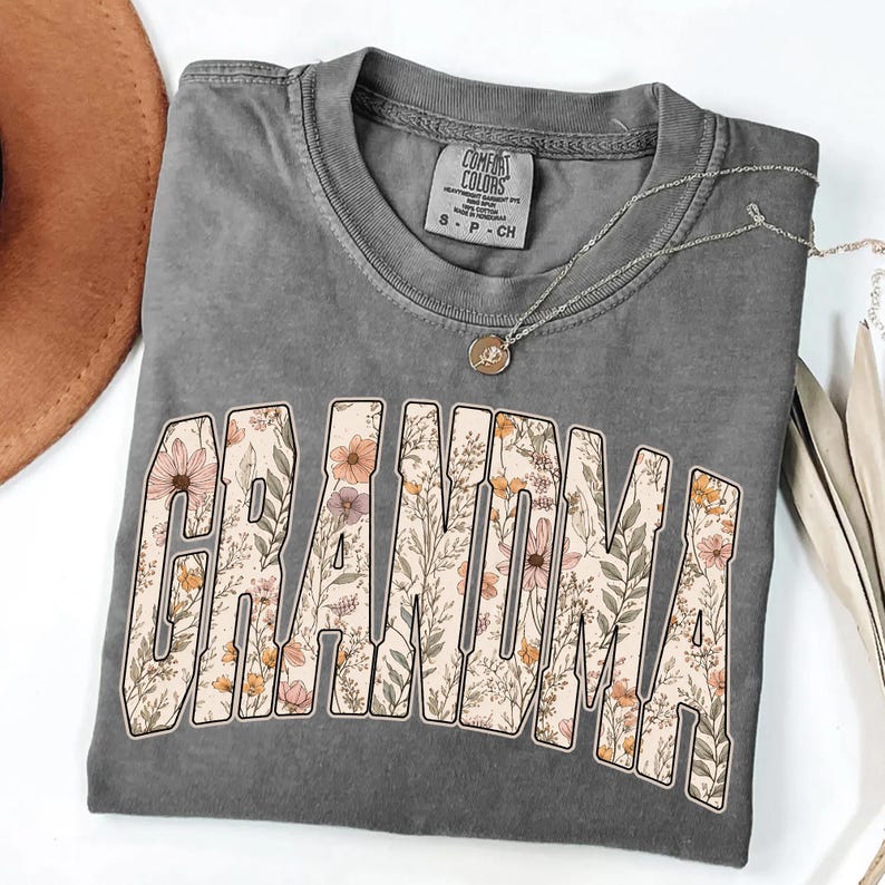 Op de afbeelding: Een grijs Comfort Colors T-shirt met het woord "GRANDMA" in een bloemenontwerp. Het shirt heeft een ronde hals en korte mouwen. Een zilveren ketting met een hanger is over het shirt gedrapeerd.
