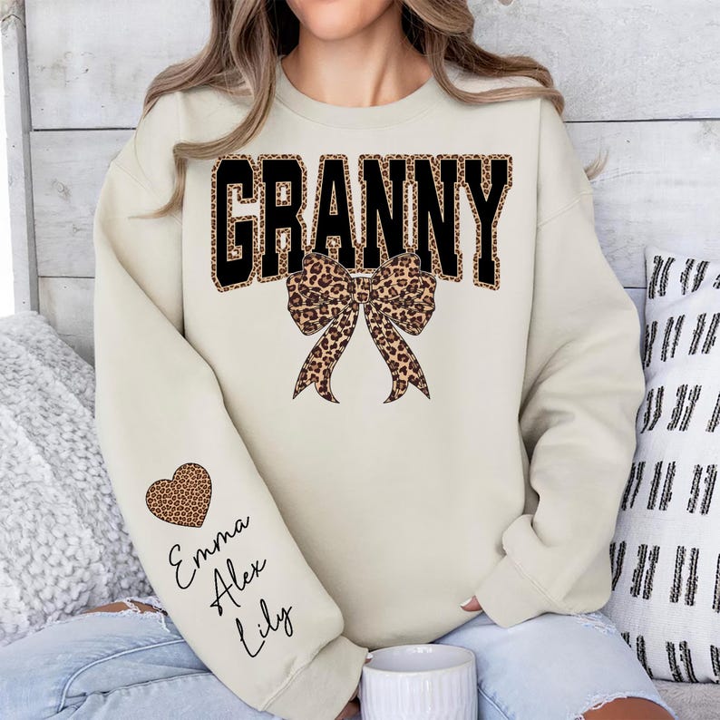 Puede incluir: Una sudadera color crema con la palabra "GRANNY" en letras negras con estampado de leopardo. Debajo de la palabra hay un lazo con estampado de leopardo. La manga izquierda tiene un coraz&oacute;n con estampado de leopardo con los nombres Emma, Alex y Lily.