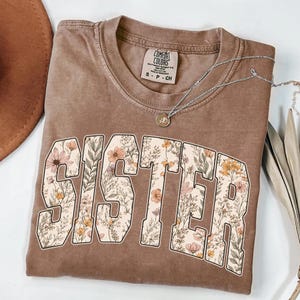 May include: A brown Comfort Colors t-shirt with the word "SISTER" in large, floral-filled letters. The shirt has a crew neck and short sleeves. A silver necklace with a small pendant is draped across the shirt.