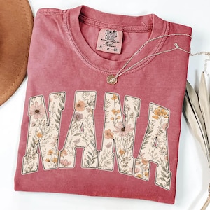 May include: A dusty rose t-shirt with the word "NANA" in large, floral-filled letters. The shirt has a crew neck and short sleeves. A silver necklace with a pendant rests on the shirt. The shirt is folded and has a tag that says "Comfort Colors".