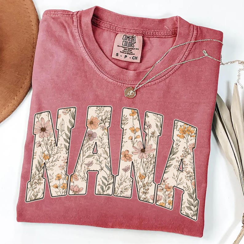 May include: A dusty rose t-shirt with the word "NANA" in large, floral-filled letters. The shirt has a crew neck and short sleeves. A silver necklace with a pendant rests on the shirt. The shirt is folded and has a tag that says "Comfort Colors".
