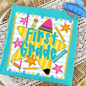 May include: A turquoise t-shirt featuring a graphic print with the text First Grade in teal letters. The design includes a large yellow star, a pencil, a glue stick, a blue ruler, and colourful stars on a background of light blue horizontal stripes.