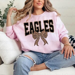 May include: A pale pink sweatshirt featuring the word "EAGLES" in black block letters with a leopard print design. A leopard print bow is positioned below the word. The sweatshirt is paired with blue jeans.