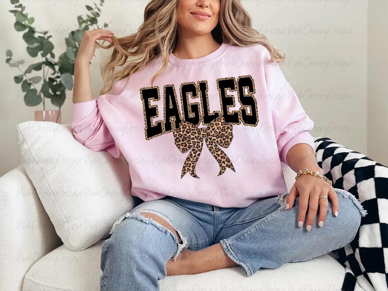 May include: A pale pink sweatshirt featuring the word "EAGLES" in black block letters with a leopard print design. A leopard print bow is positioned below the word. The sweatshirt is paired with blue jeans.