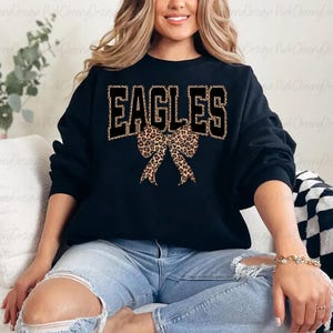 May include: Black sweatshirt featuring the word "EAGLES" in black block letters with a leopard print fill. A leopard print bow is centered below the word. The sweatshirt is worn with blue jeans.