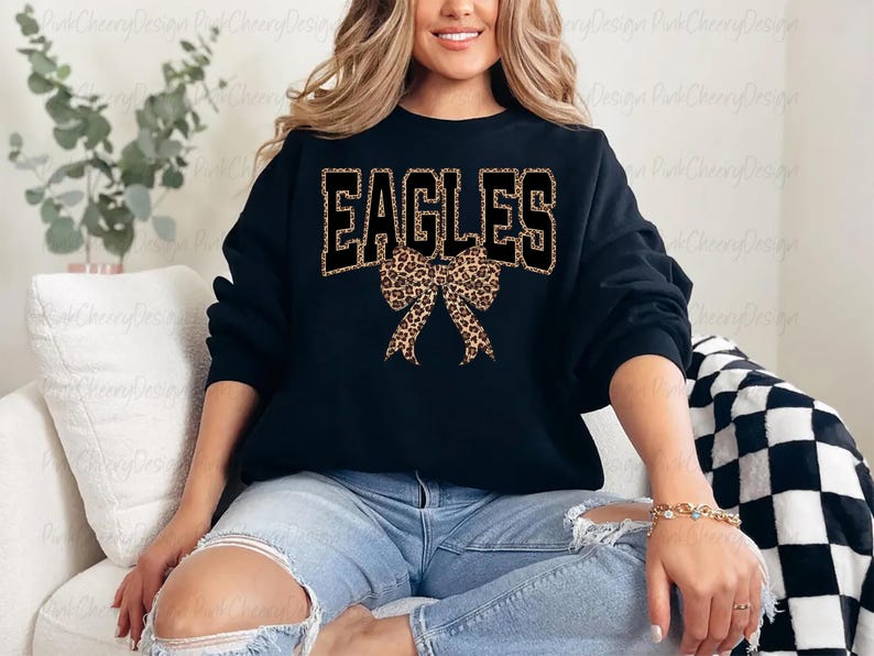 May include: Black sweatshirt featuring the word "EAGLES" in black block letters with a leopard print fill. A leopard print bow is centered below the word. The sweatshirt is worn with blue jeans.