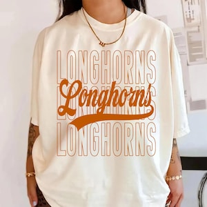May include: Cream-colored t-shirt with the word "LONGHORNS" in a stacked, orange retro font. The shirt has short sleeves and a relaxed fit, suitable for casual wear.
