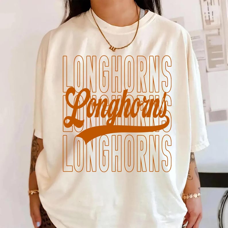 Puede incluir: Camiseta color crema con la palabra "LONGHORNS" en una fuente retro naranja apilada. La camiseta tiene mangas cortas y un corte relajado, adecuada para uso casual.