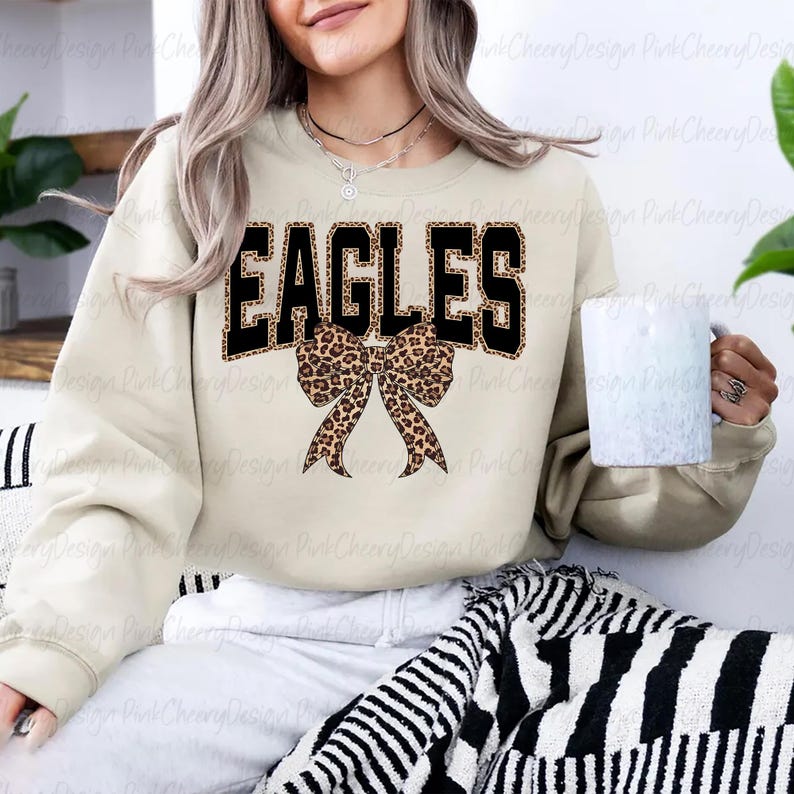 May include: A cream-coloured sweatshirt featuring the word "EAGLES" in black block letters with a leopard print trim. A leopard print bow is positioned below the word. A white mug is held.