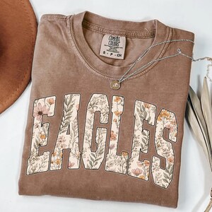 Puede incluir: Una camiseta marr&oacute;n con la palabra "EAGLES" en un dise&ntilde;o floral. La camiseta tiene cuello redondo y mangas cortas. Un collar plateado con un peque&ntilde;o colgante est&aacute; drapeado sobre la camiseta. La camiseta tiene una etiqueta Comfort Colors.