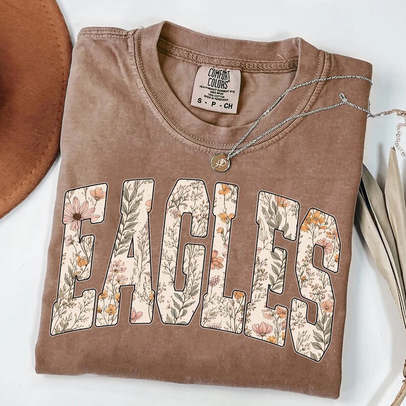 Puede incluir: Una camiseta marr&oacute;n con la palabra "EAGLES" en un dise&ntilde;o floral. La camiseta tiene cuello redondo y mangas cortas. Un collar plateado con un peque&ntilde;o colgante est&aacute; drapeado sobre la camiseta. La camiseta tiene una etiqueta Comfort Colors.