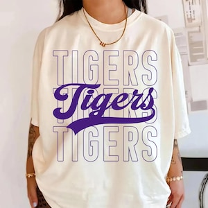 Könnte beinhalten: Cremefarbenes T-Shirt mit dem Wort "TIGERS" dreifach in einem gestapelten Design bedruckt. Die oberen und unteren Instanzen sind lila umrandet, während das mittlere "Tigers" durchgehend lila ist.