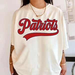 Pode incluir: Camiseta creme com a palavra "Patriots" em escrita vermelha com contorno azul. A palavra está acima de uma faixa vermelha curva. A camisa tem mangas curtas e um corte folgado.