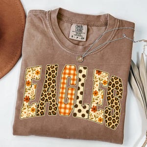Peut inclure: Un t-shirt marron à manches courtes avec le mot EAGLES imprimé sur la poitrine. Chaque lettre présente un motif différent, notamment des imprimés floraux, des taches de léopard, du vichy orange et des pois marron. Étiquette Comfort Colors.