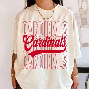 Puede incluir: Camiseta color crema con la palabra "CARDINALS" en rojo, repetida tres veces. La palabra "Cardinals" del medio est&aacute; en cursiva con una l&iacute;nea roja debajo. Una camiseta informal de manga corta.
