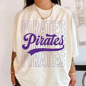 May include: Cream-colored t-shirt with the word "PIRATES" stacked three times in a purple outline font. The word "Pirates" is in a cursive purple font in the middle. The shirt is a casual style.