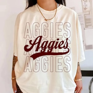 May include: Cream-colored t-shirt with the word "AGGIES" in a stacked design. The top and bottom "AGGIES" are outlined in a dark red, while the middle "Aggies" is in a script font with a dark red banner.
