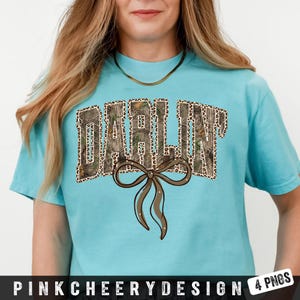 May include: A light blue t-shirt with the word "DARLIN'" in a camouflage and leopard print design. A brown bow with rhinestones is below the text. The shirt has short sleeves and a crew neck.