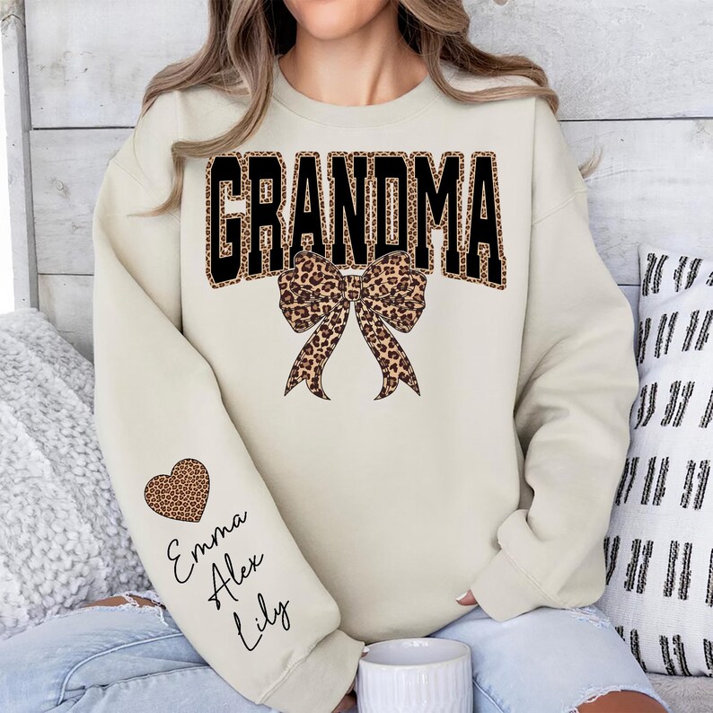 Puede incluir: Una sudadera color crema con la palabra "GRANDMA" en letras negras con estampado de leopardo. Un lazo con estampado de leopardo est&aacute; centrado debajo de la palabra. La manga izquierda tiene un coraz&oacute;n con estampado de leopardo con los nombres "Emma, Alex, Lily" en cursiva.