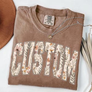 May include: A brown Comfort Colors t-shirt with the word "CUSTOM" in large, floral-filled letters. The shirt has a crew neck and short sleeves. A silver necklace with a small pendant is draped across the shirt.