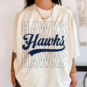 May include: Cream-colored t-shirt with the word "Hawks" in a layered design. The top and bottom "Hawks" are outlined in a navy blue, while the middle "Hawks" is solid navy blue with a curved underline. The shirt has short sleeves.