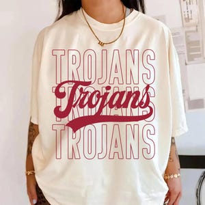 May include: Cream-colored t-shirt with the word "TROJANS" in a stacked design. The top and bottom text are outlined in red, while the middle text is solid red. The shirt has short sleeves and a relaxed fit.